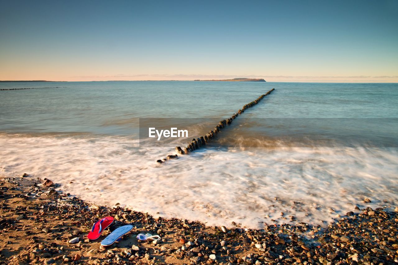 Flip flops and white swimming goggles ready on stony beach at wooden breakwater. wavy sea level.