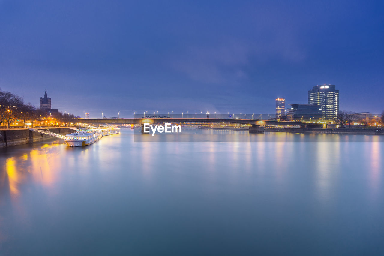 Illuminated bridge over river by buildings against sky at dusk