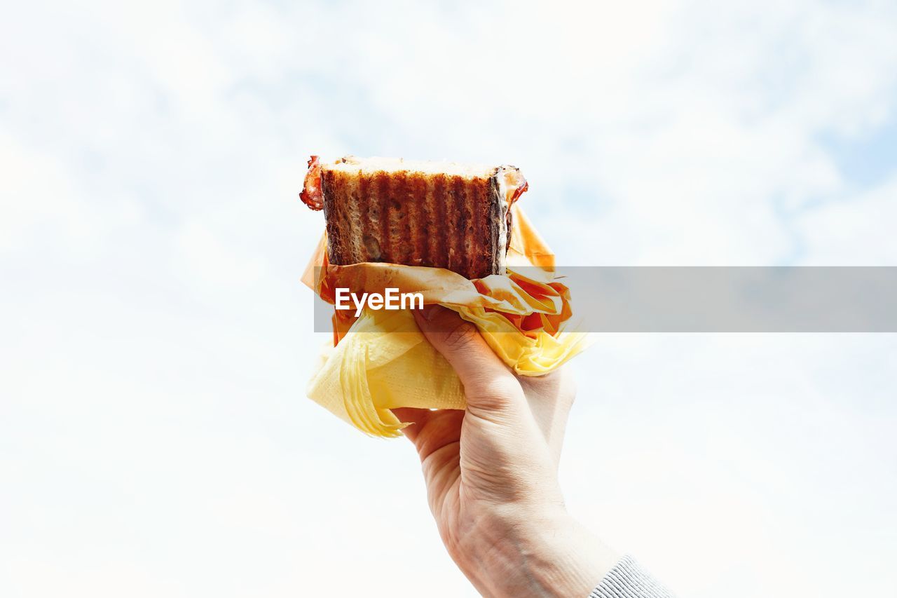 Cropped hand holding sandwich against cloudy sky