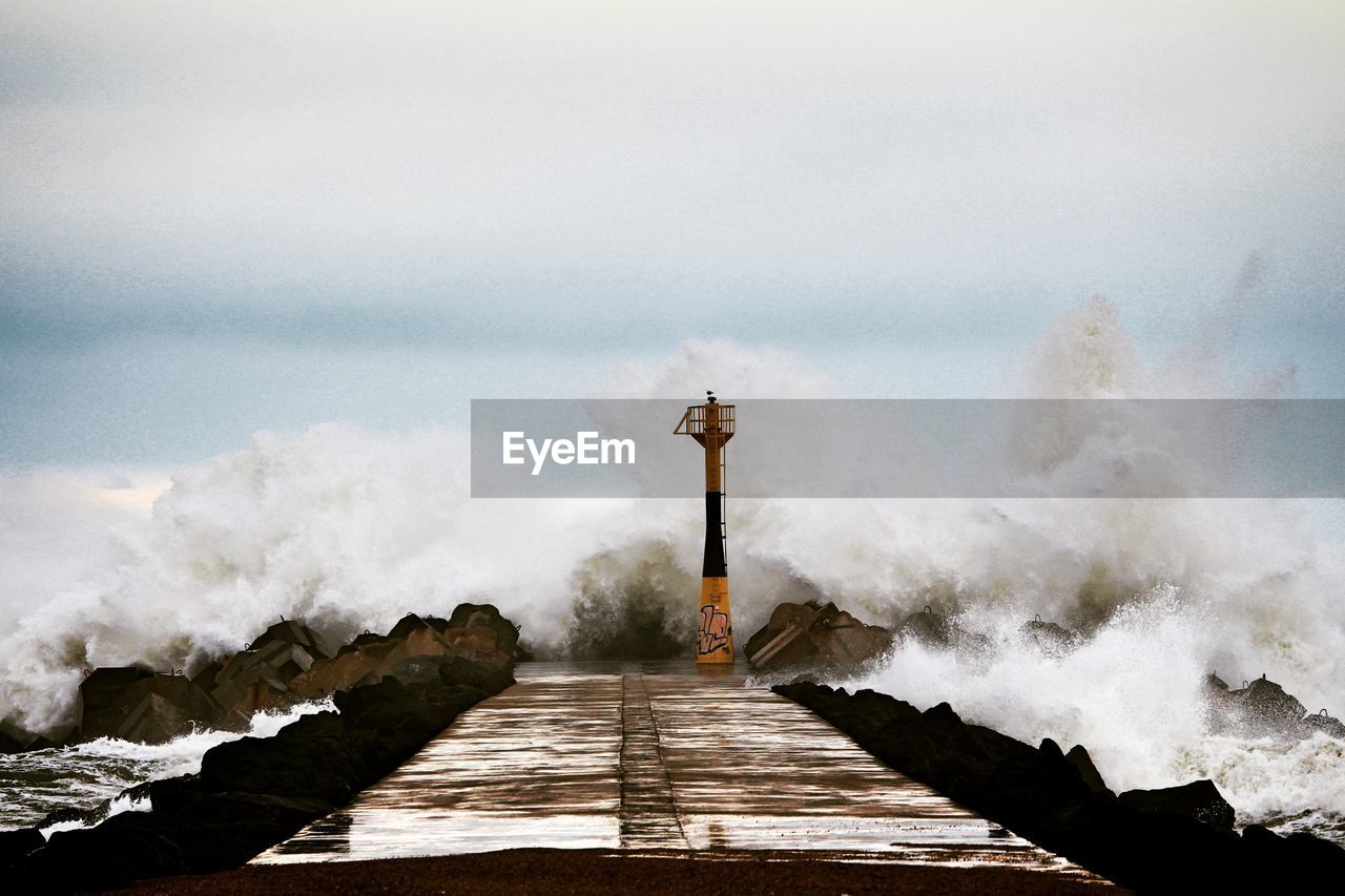Waves splashing by lighthouse and pier against cloudy sky during sunset