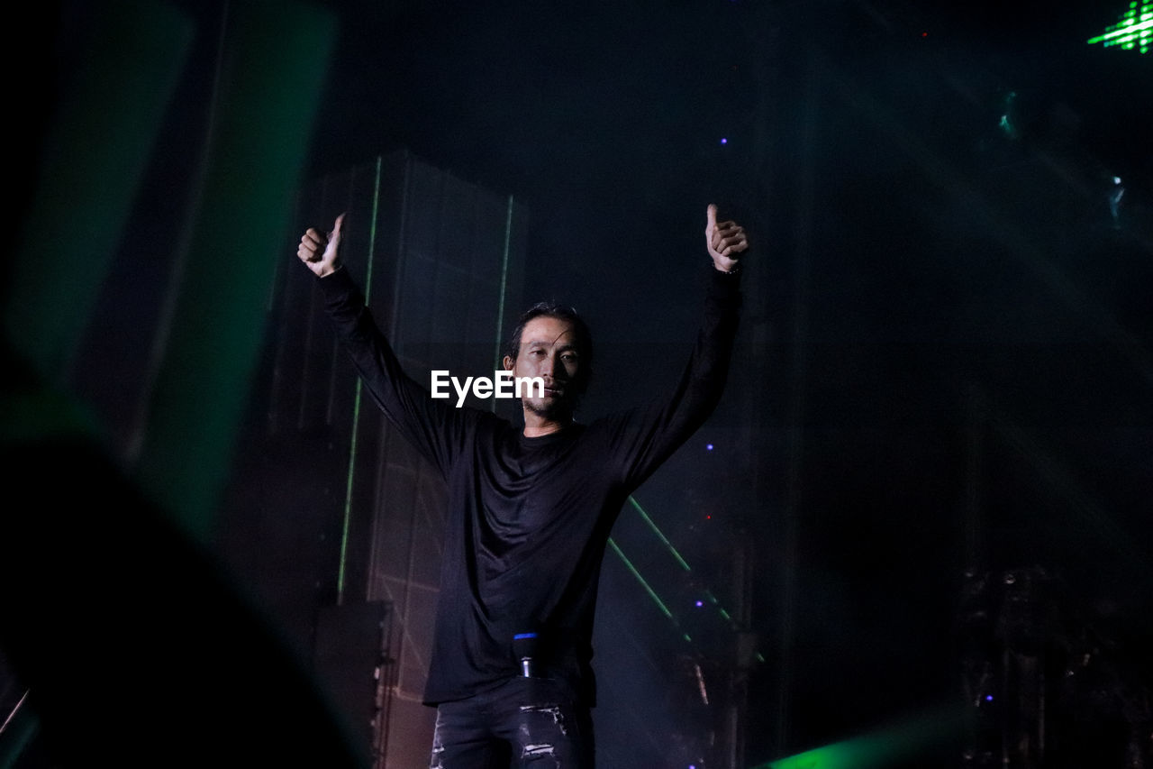 Portrait of young man with arms raised showing thumbs up while standing on stage at night