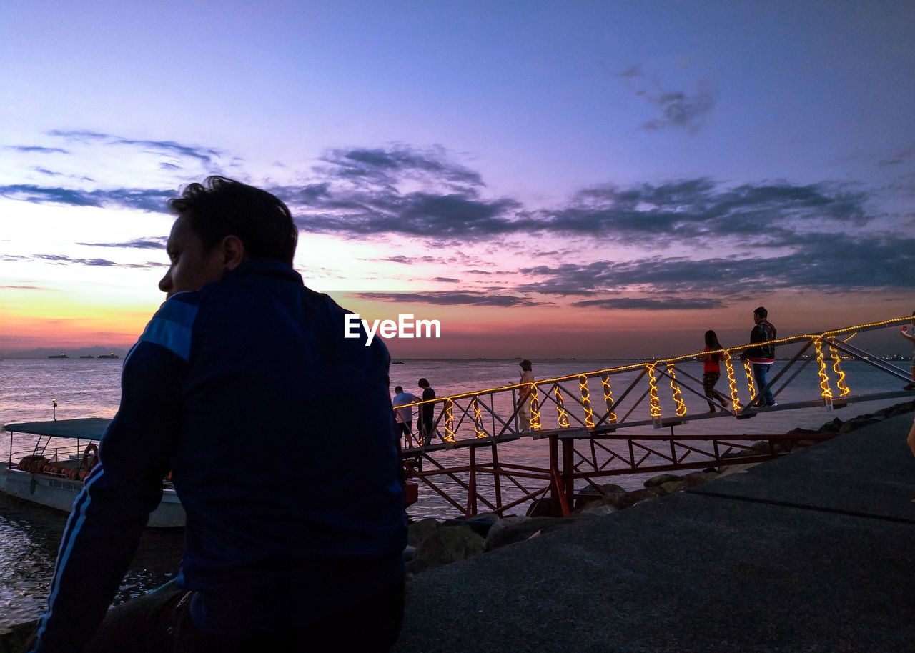 MAN STANDING ON RAILING AGAINST SEA AT SUNSET