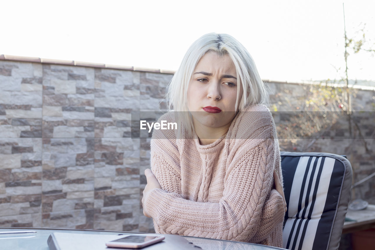 Sad young woman sitting at table against wall