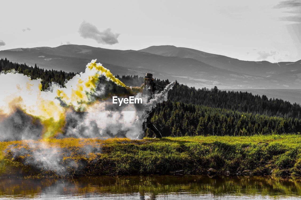 Woman holding distress flare while standing on grass by lake against mountains and sky