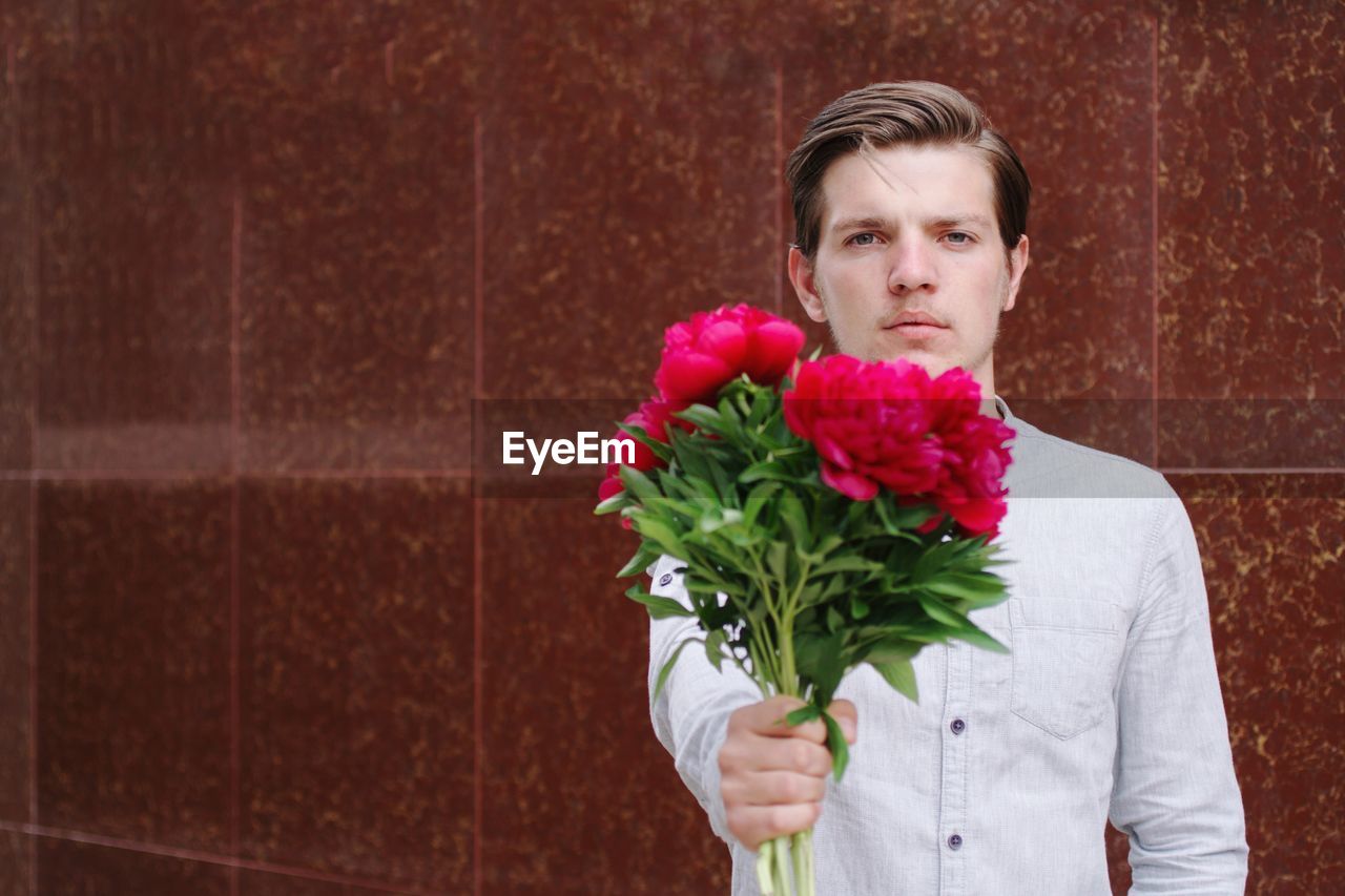 Portrait of handsome man standing against red wall holding flower in hand