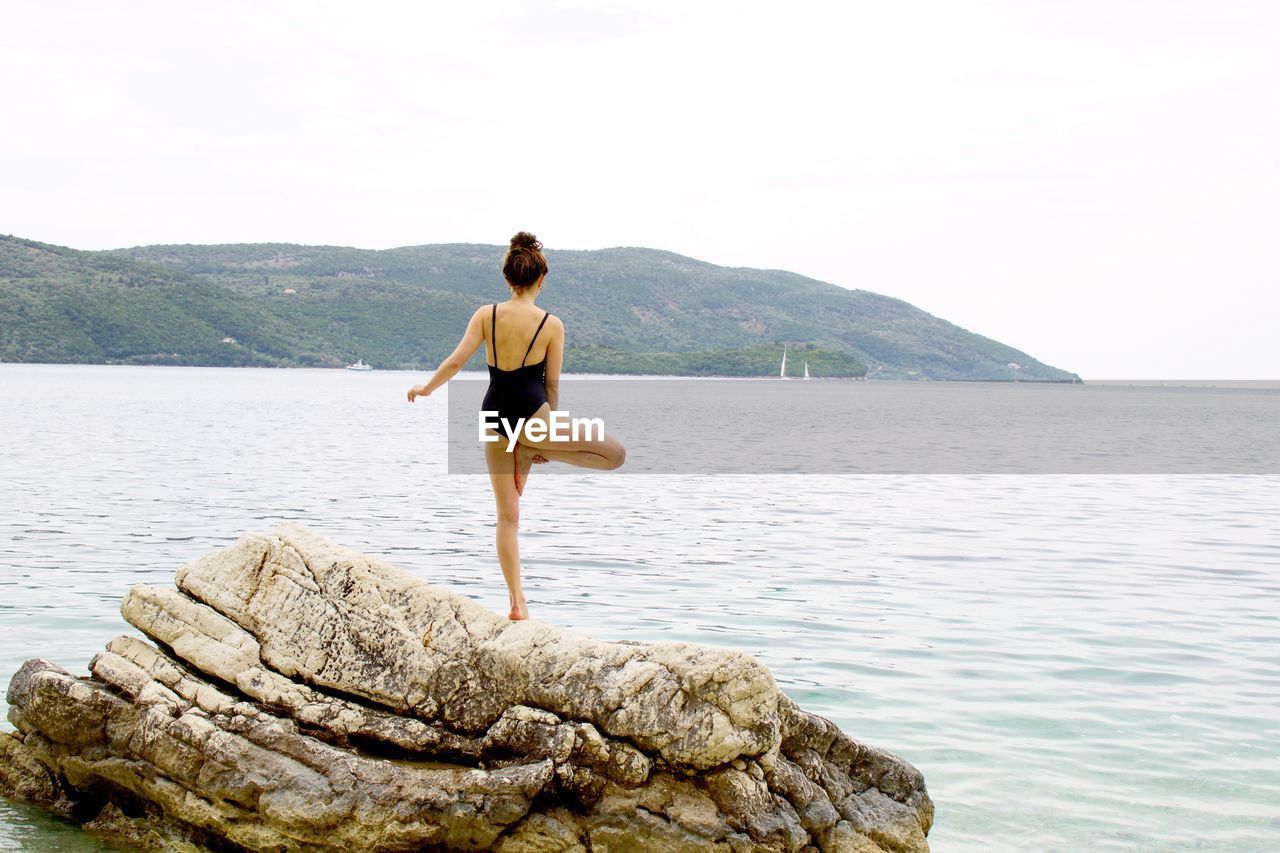 Rear view of woman in swimsuit doing yoga on rock by sea against sky