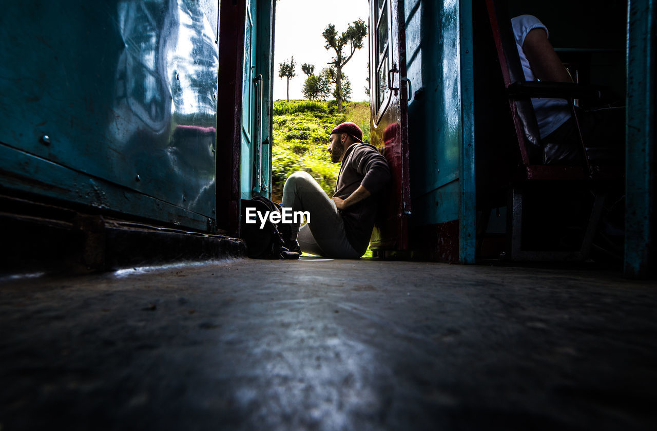 Side view of male traveler sitting in doorway of aged cabin while enjoying green trees during trip in sri lanka