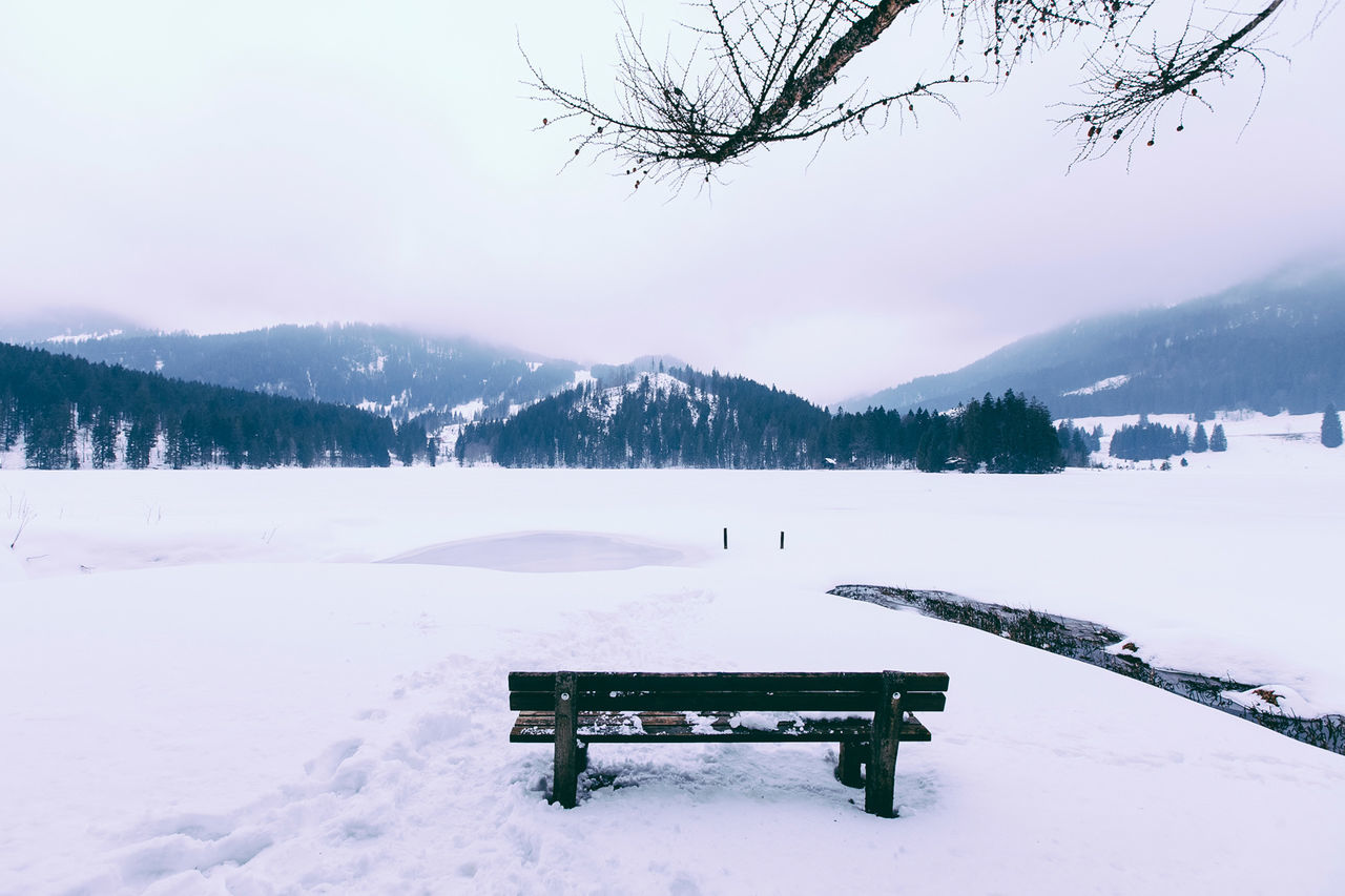 Wooden bench on snow covered field against sky