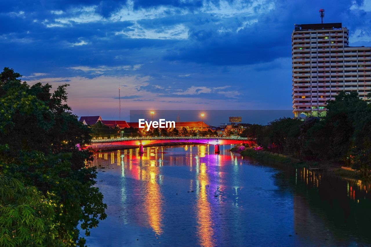 ILLUMINATED BUILDINGS BY RIVER AGAINST SKY