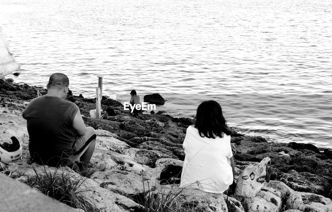 REAR VIEW OF PEOPLE SITTING ON ROCK AT SHORE