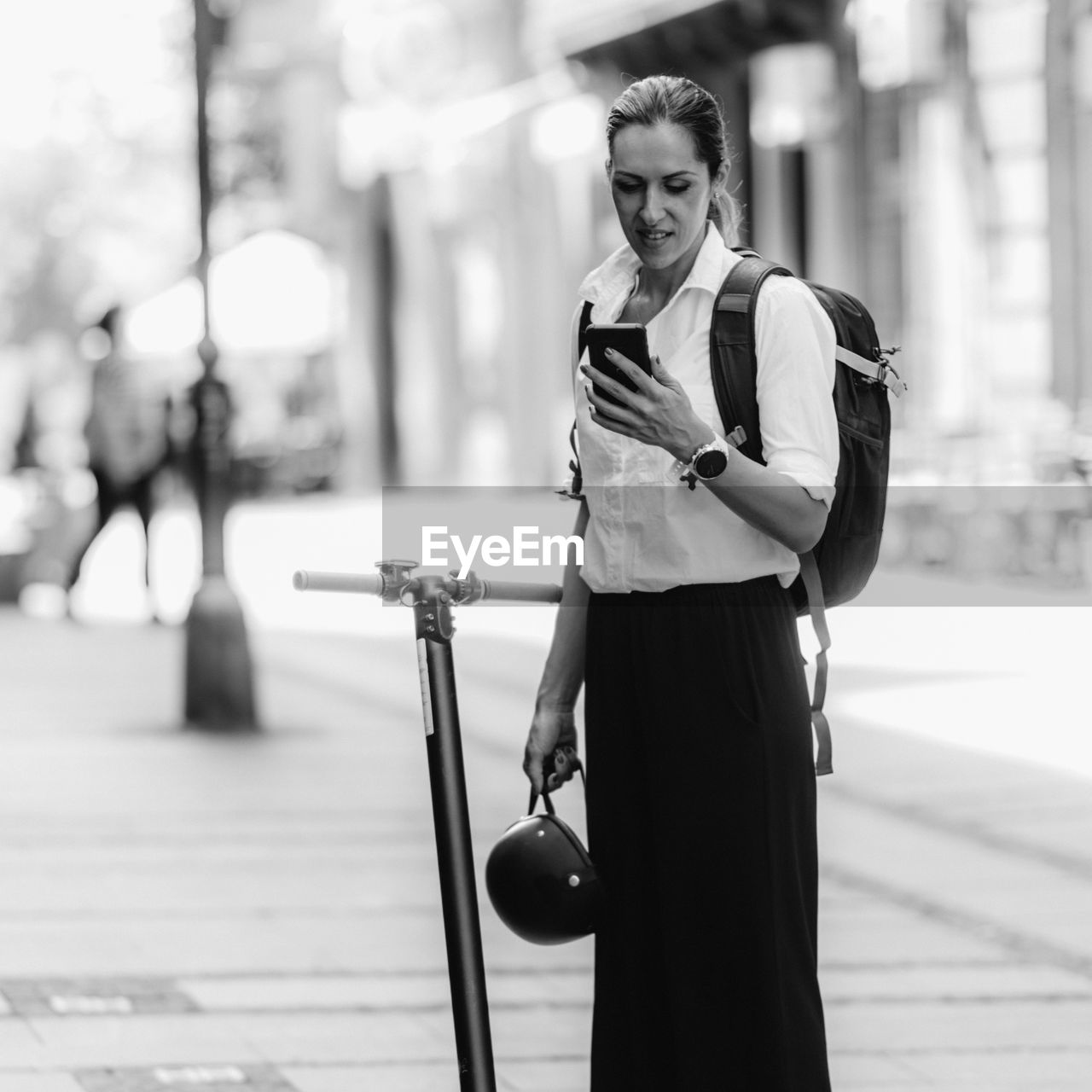 Businesswoman standing and using her smart phone with electric scooter next to her in the city.