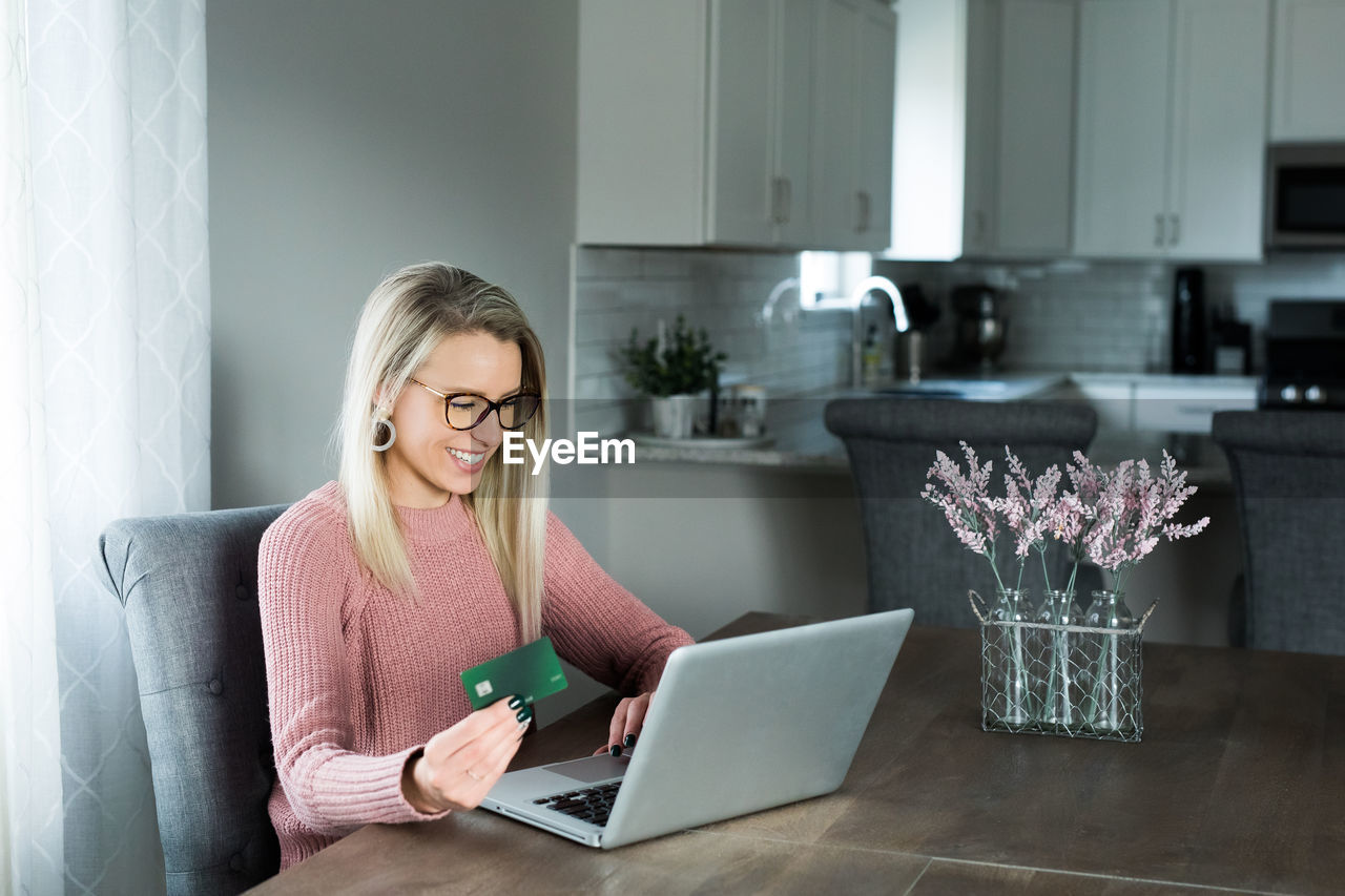 Happy young woman holding credit card while using laptop at home