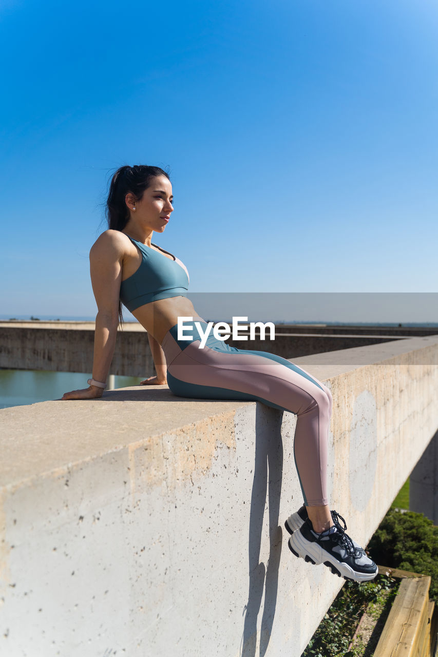 Side view of fit female athlete in sports clothes looking away from cement fence under blue sky