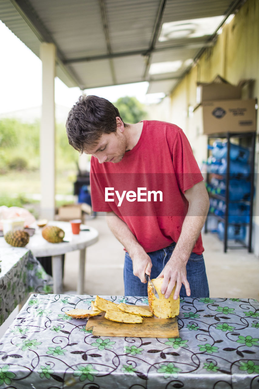 Young man cutting fresh, organic pineapple on wood cutting board.