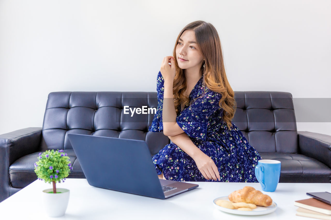 YOUNG WOMAN USING LAPTOP WHILE SITTING ON SOFA