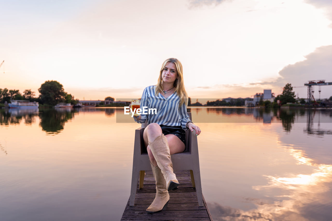 water, reflection, sky, one person, adult, nature, lake, women, relaxation, sitting, sunset, young adult, evening, beauty in nature, smiling, lifestyles, casual clothing, happiness, emotion, leisure activity, full length, tranquility, fashion, blond hair, architecture, hairstyle, cloud, summer, copy space, outdoors, trip, long hair, holiday, clothing, vacation, travel destinations, twilight, travel, striped, sunlight, dusk, female, tranquil scene, portrait, pier, city, front view