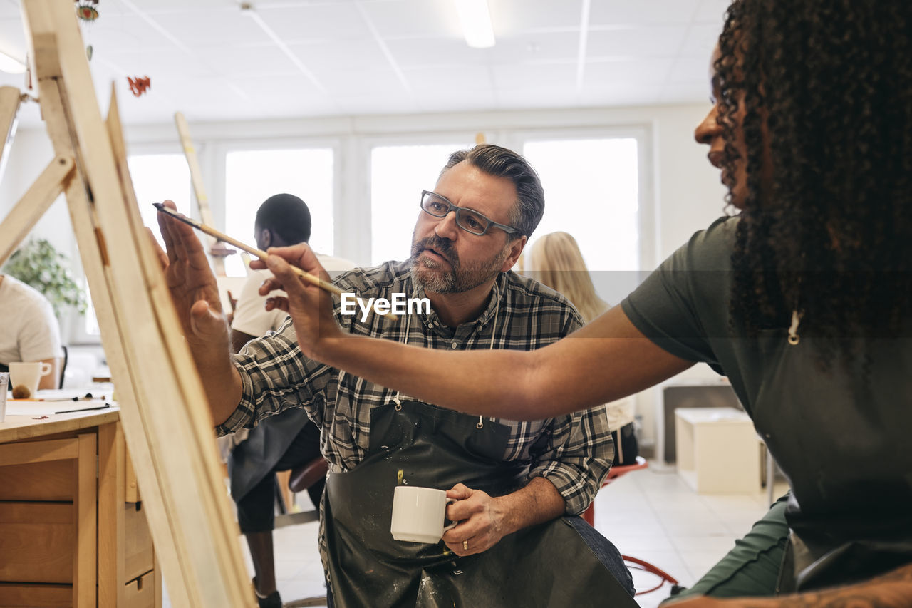 Male tutor assisting female student in painting on artist's canvas in art class