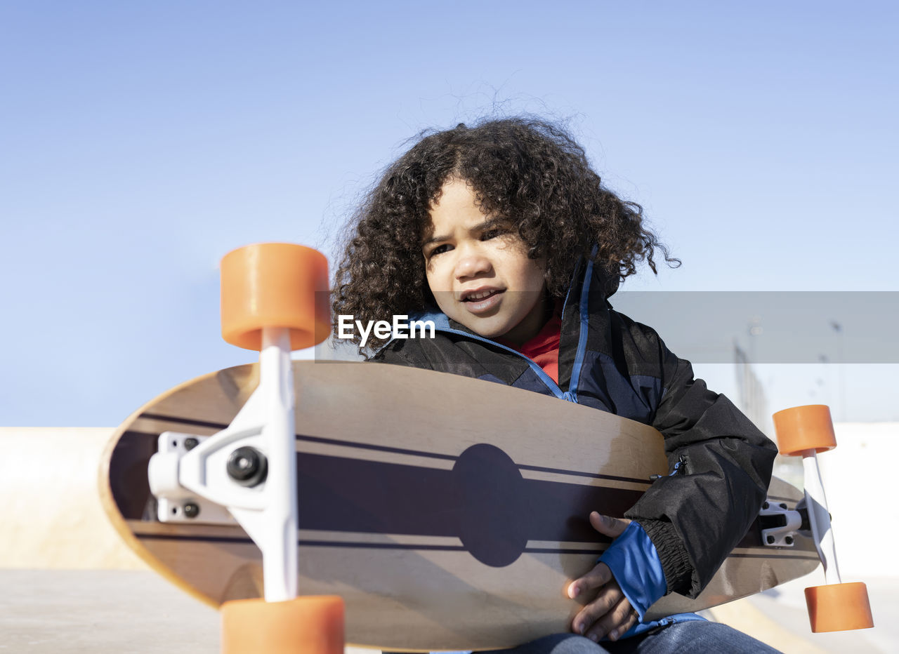 Ethnic child with curly hair sitting on ramp with longboard in skate park and looking away