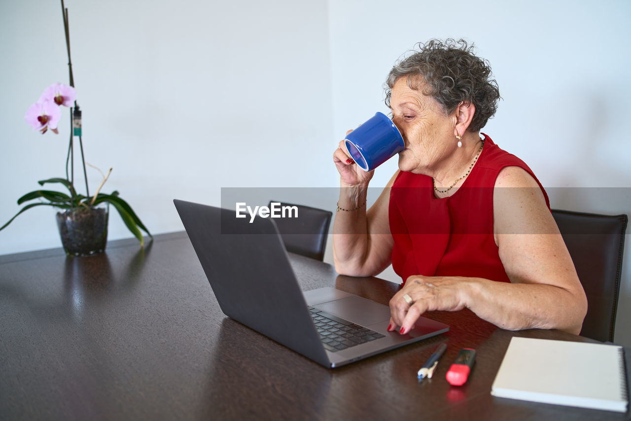 Senior woman drinks hot beverage in blue mug during video call on her laptop.