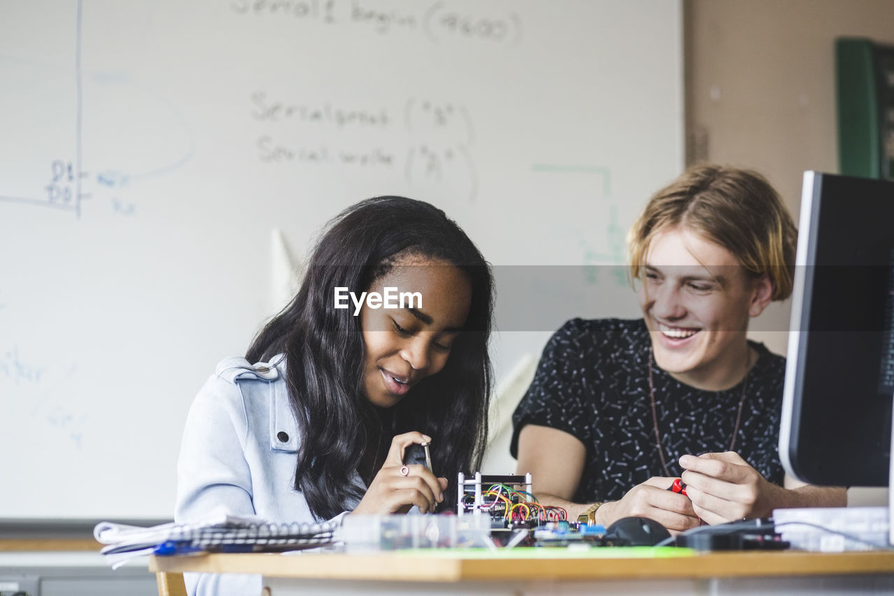 Smiling female student preparing science project with young male friend on desk in classroom at high school