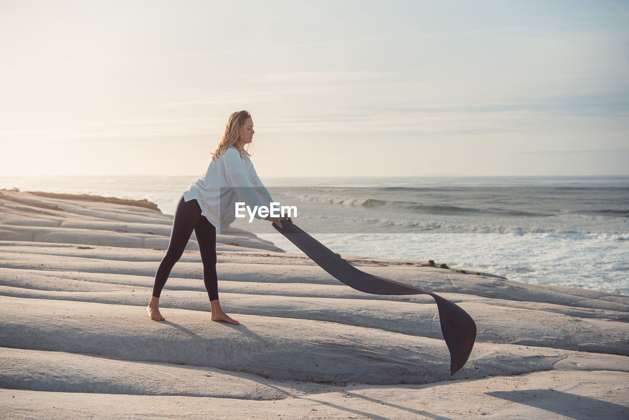 Full length of woman holding exercise mat on beach during sunset