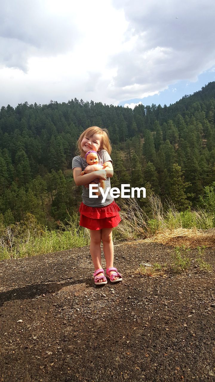 Full length of smiling girl with doll standing on footpath against trees in sunny day