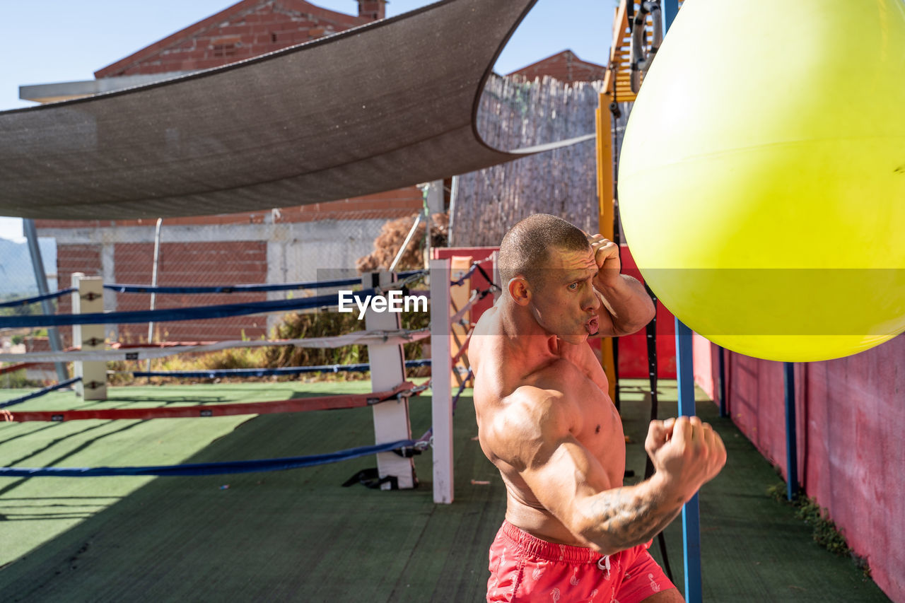 Strong sportsman with muscular body punching boxing bag while training on terrace of sports center in summer