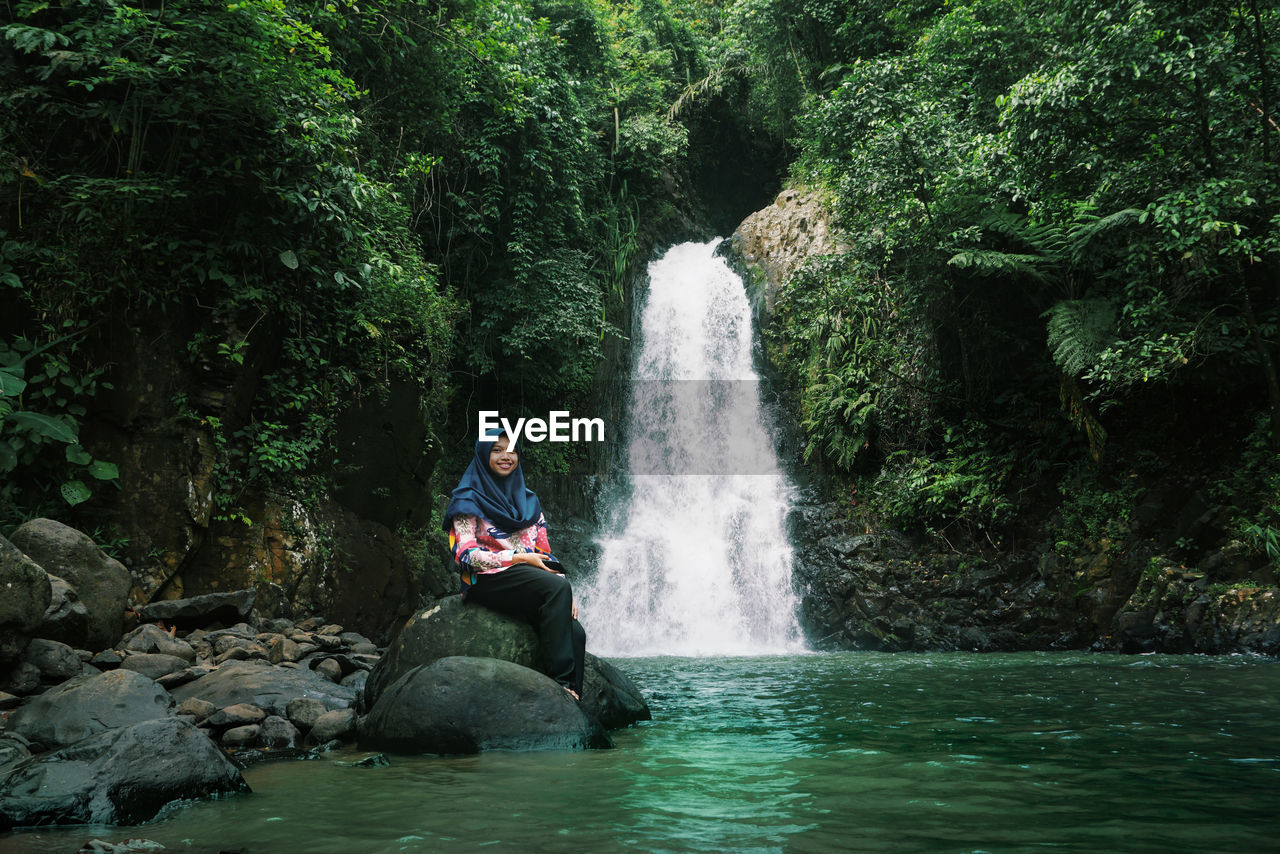 Woman sitting on rock against waterfall