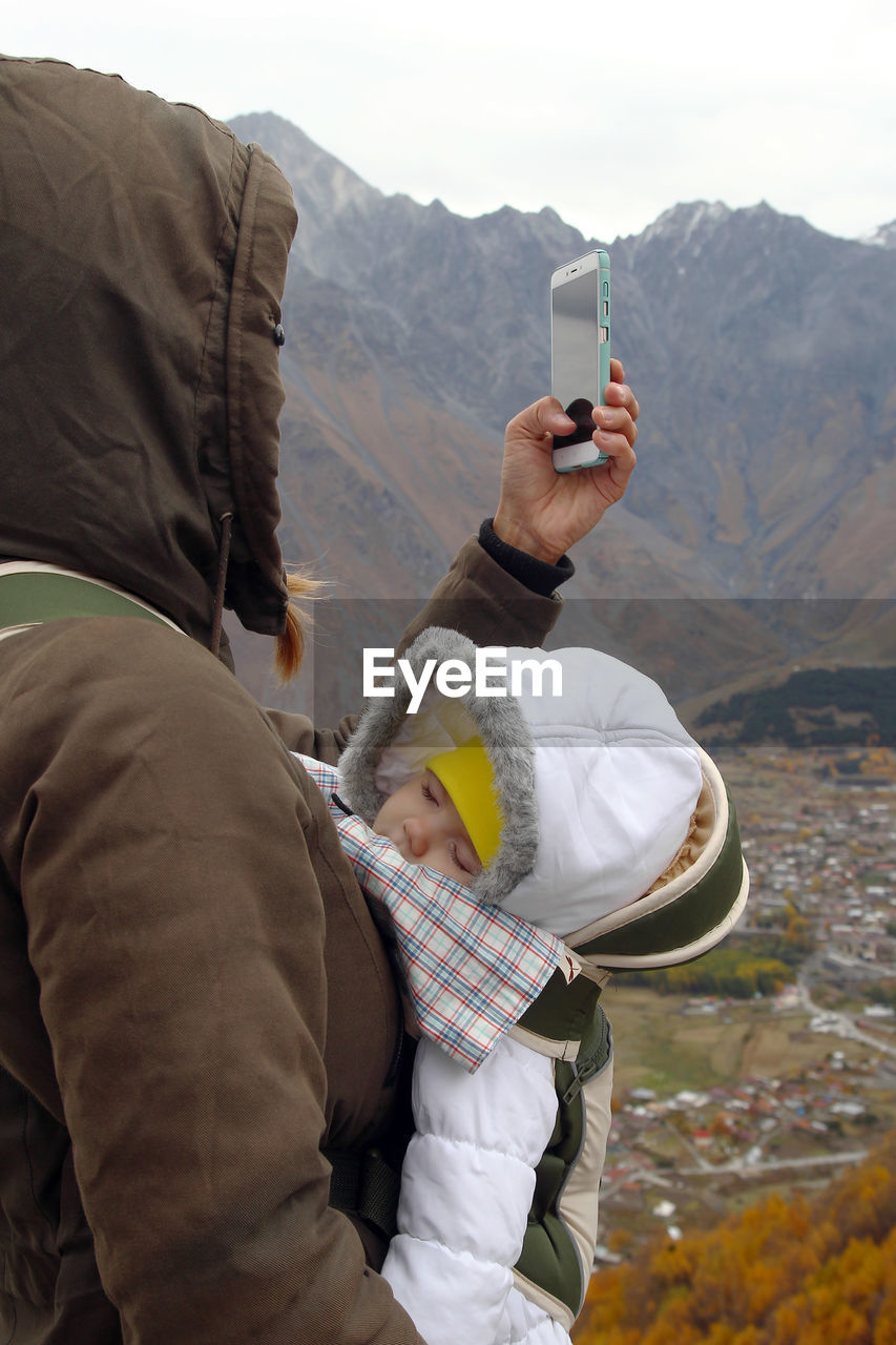 Side view of woman using phone while standing with baby against mountains