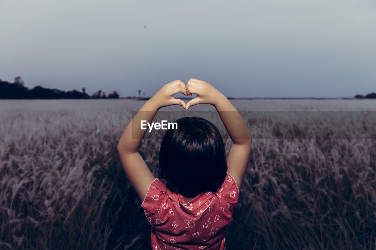 Rear view of girl making heart shape while standing on land against clear sky