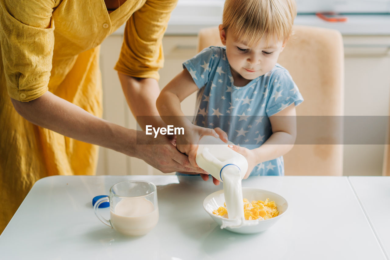 Toddler little daughter and her mother pour milk into a bowl of corn flakes.