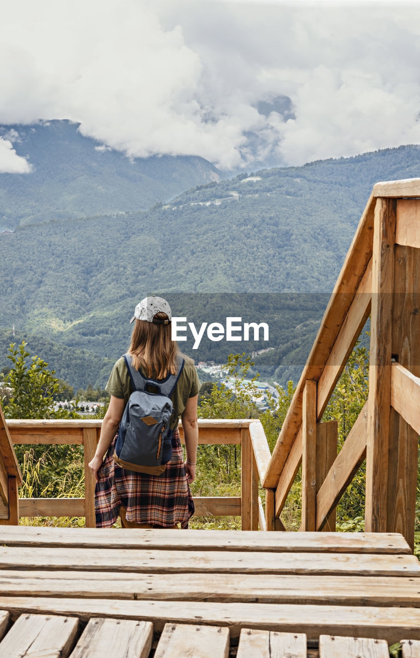 Rear view of young blonde woman  with backpack looking at view of mountains on wooden viewpoint