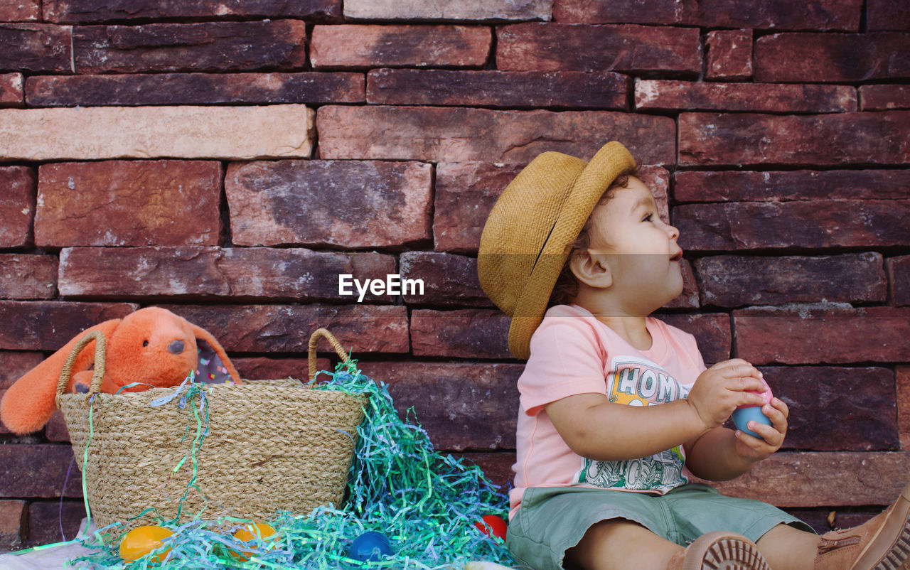 childhood, one person, child, sitting, hat, clothing, person, brick, toddler, wall, brick wall, container, fashion accessory, human face, casual clothing, wall - building feature, female, day, women, three quarter length, lifestyles, full length, cute, basket, leisure activity, innocence, outdoors, sun hat, looking, toy, nature