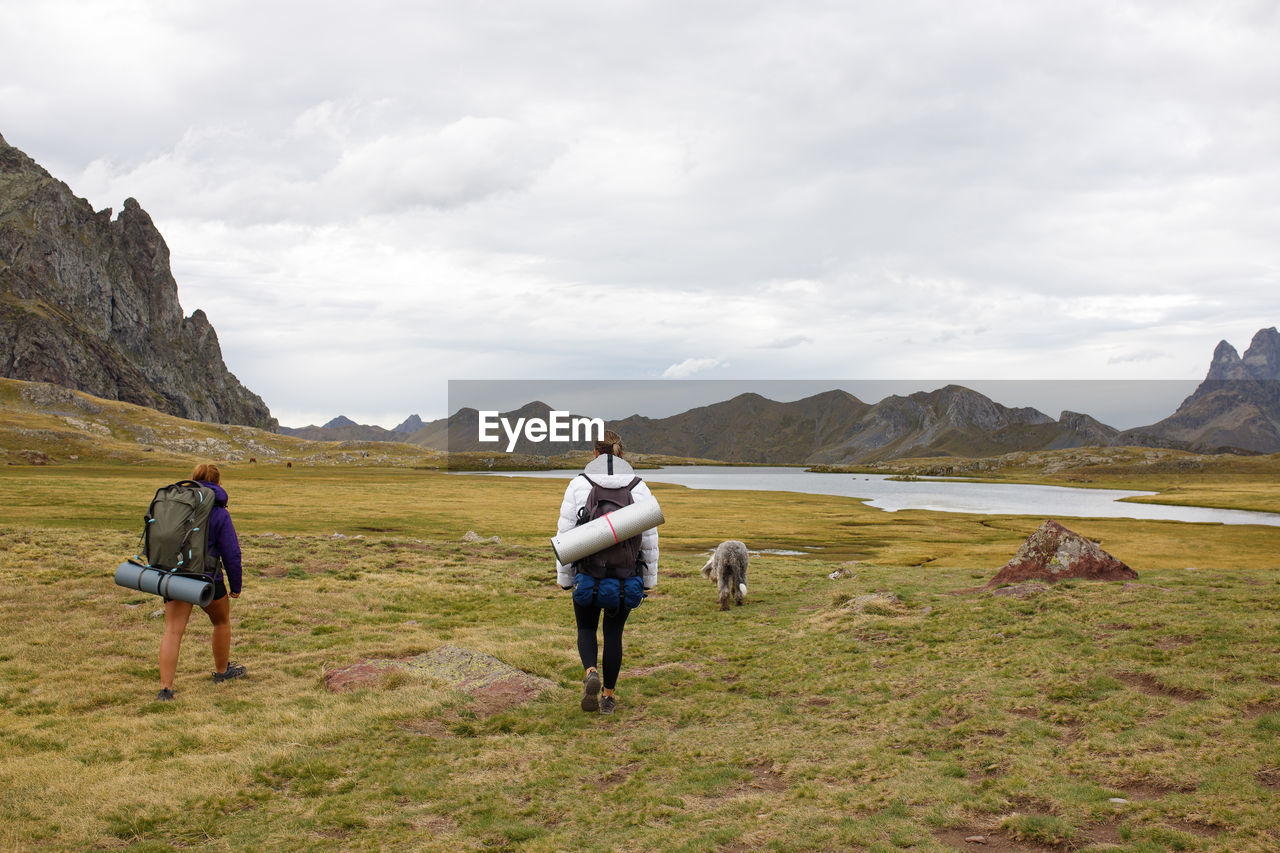 Anonymous women with dog strolling in grassy valley against mountain ridge and cloudy sky on summer day