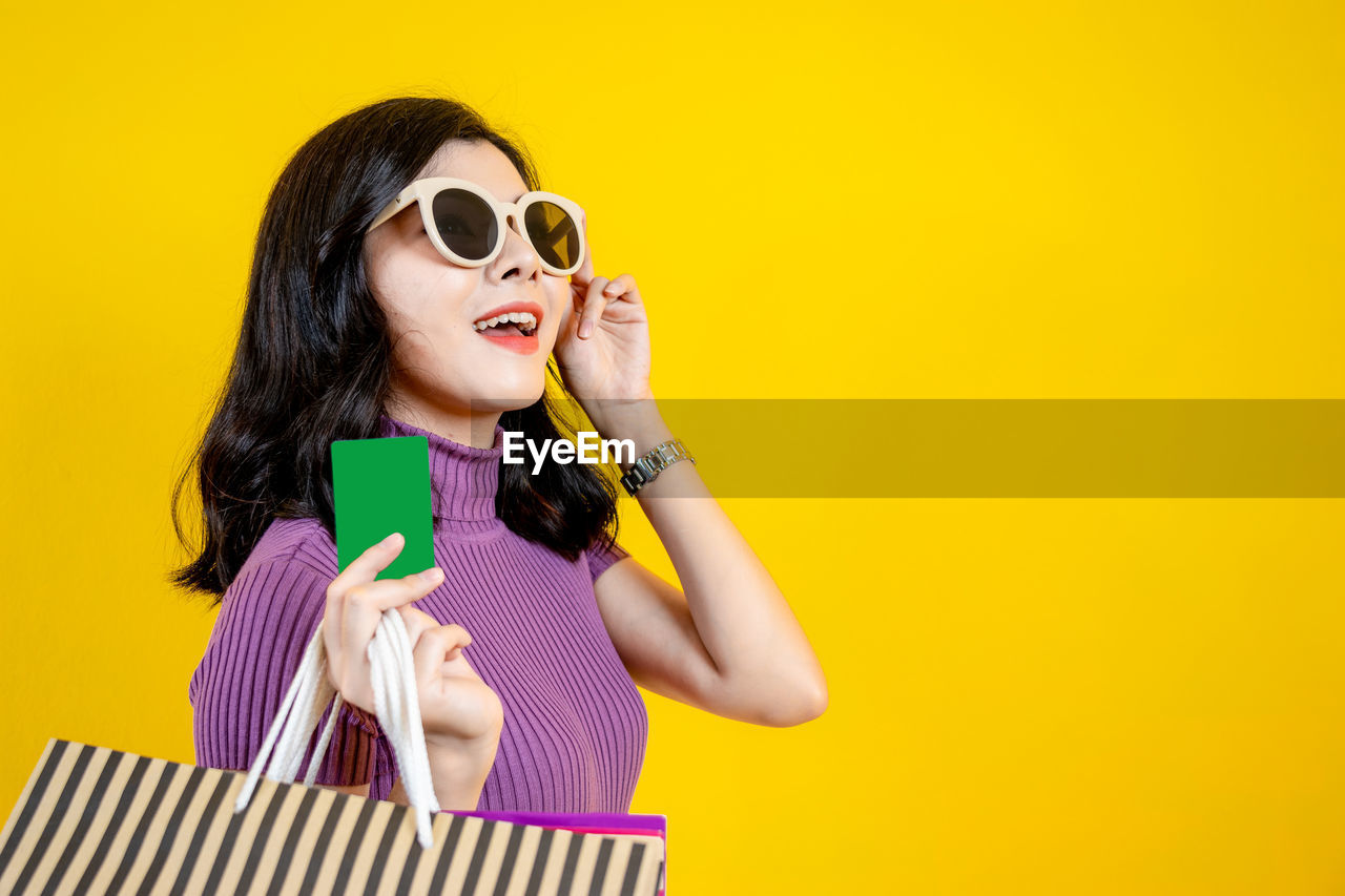 Young woman wearing sunglasses holding shopping bags while standing against yellow background