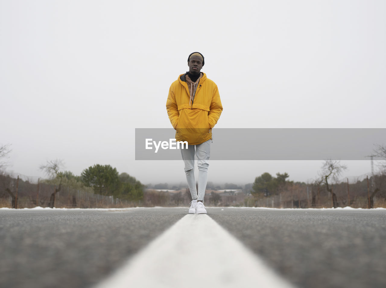 Ground level full body of african american male with hands in pockets standing on asphalt roadway among leafless trees and snow