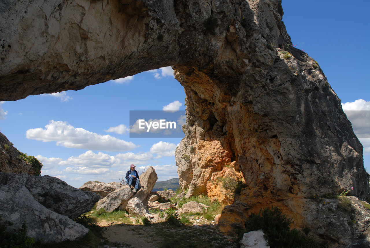 Mid distance view of man sitting at la forada rock arch against sky
