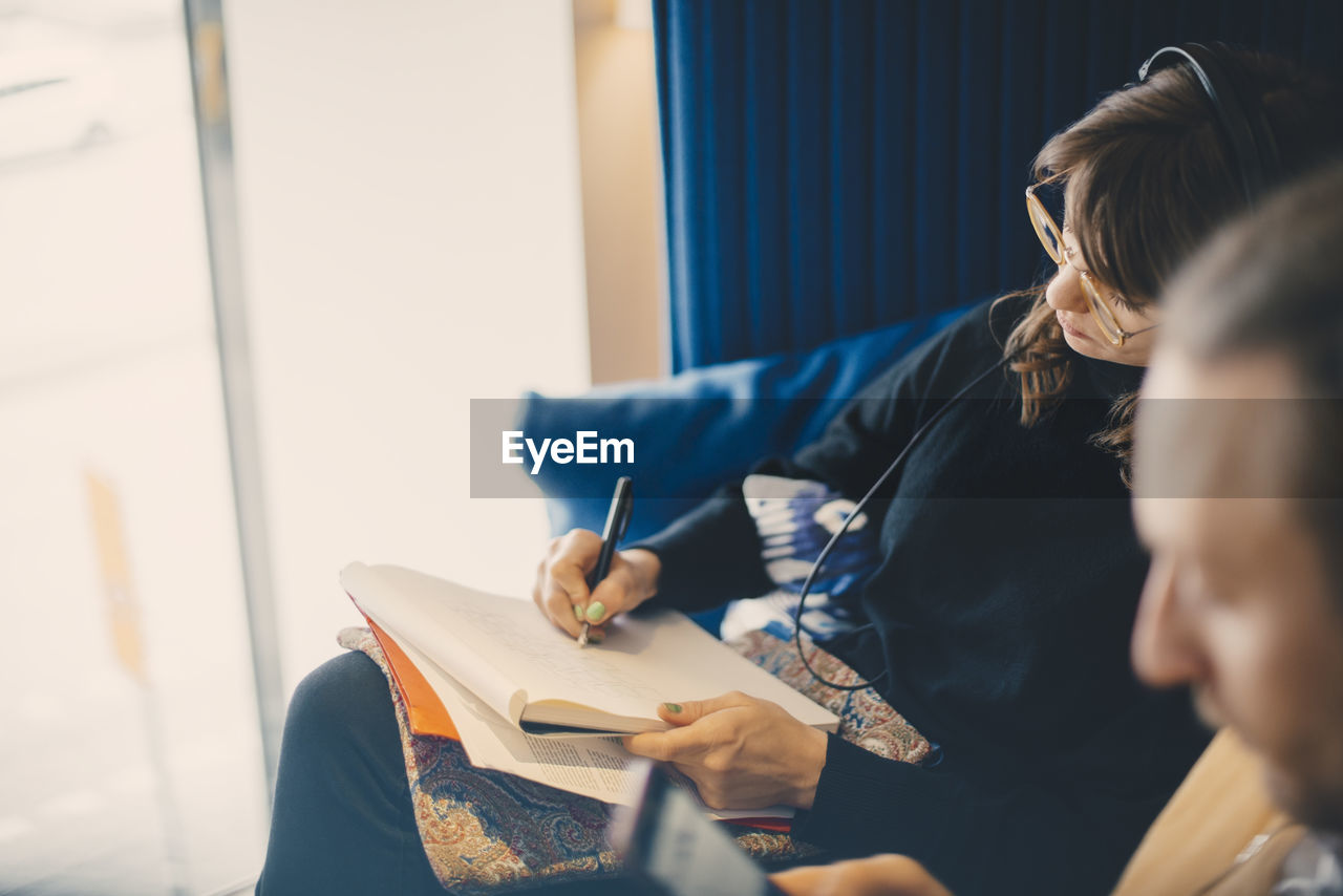 Young businesswoman listening to headphones while drawing on notepad sitting by male colleague at office cafeteria