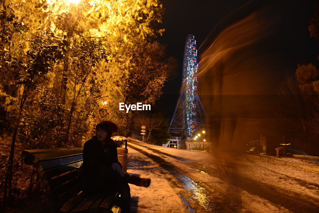 Woman sitting on bench by ghost in amusement park at night during winter