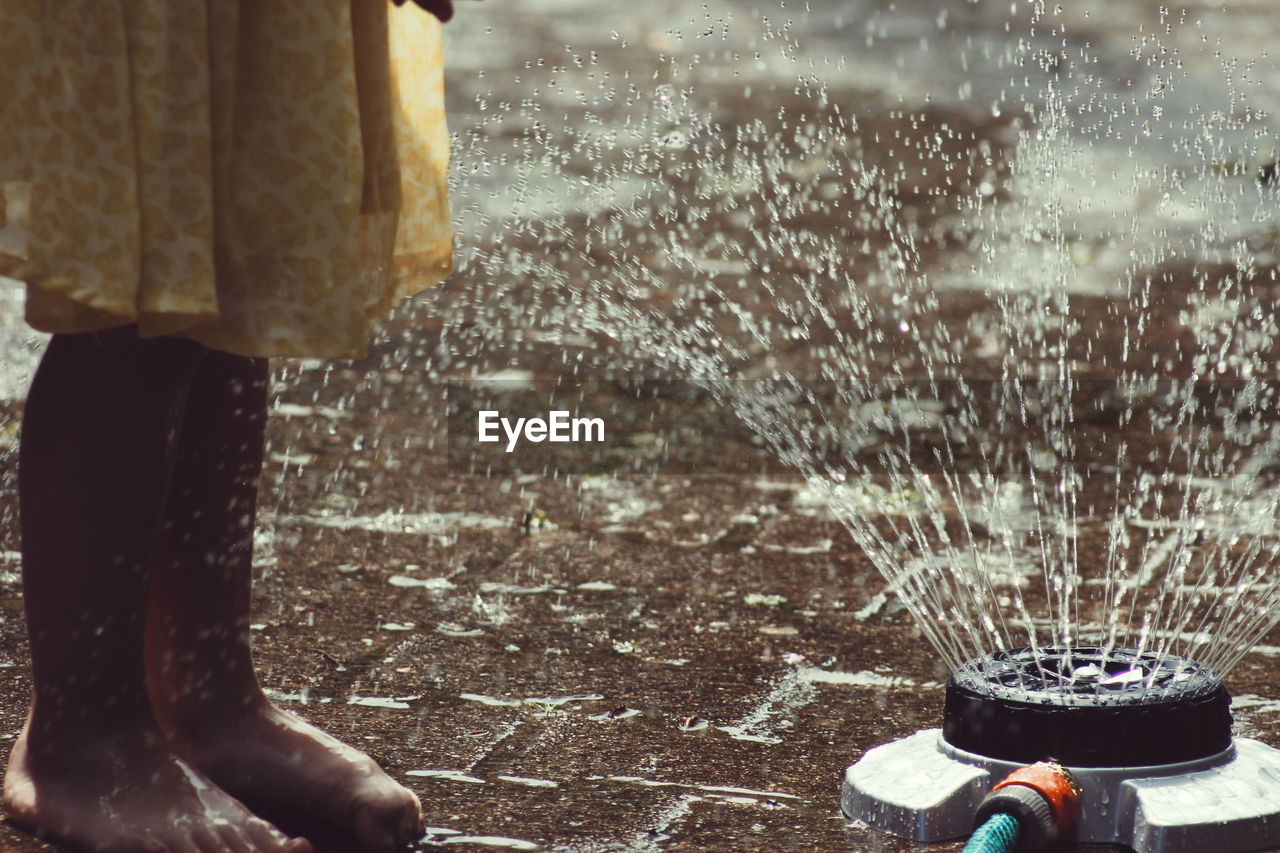 Low section of girl standing by sprinkler on footpath