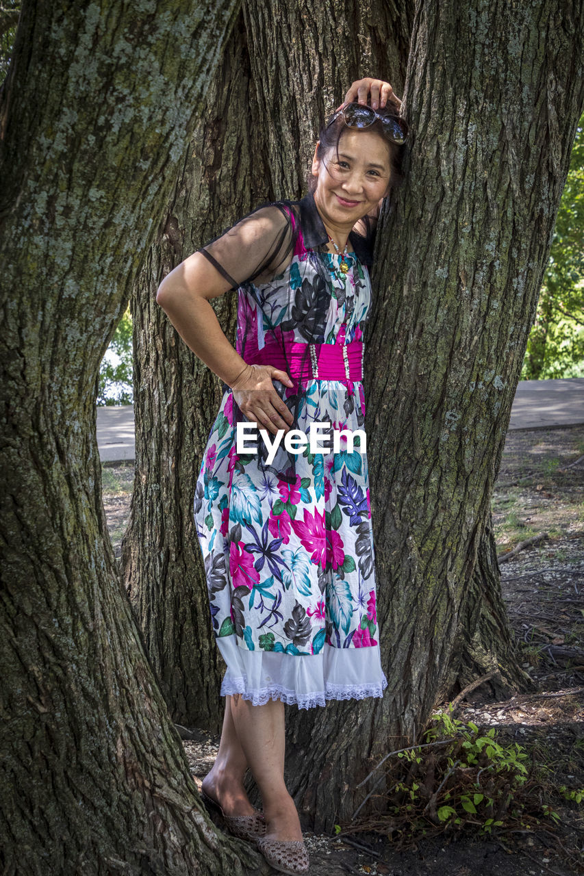 PORTRAIT OF SMILING WOMAN STANDING AGAINST TREE TRUNK