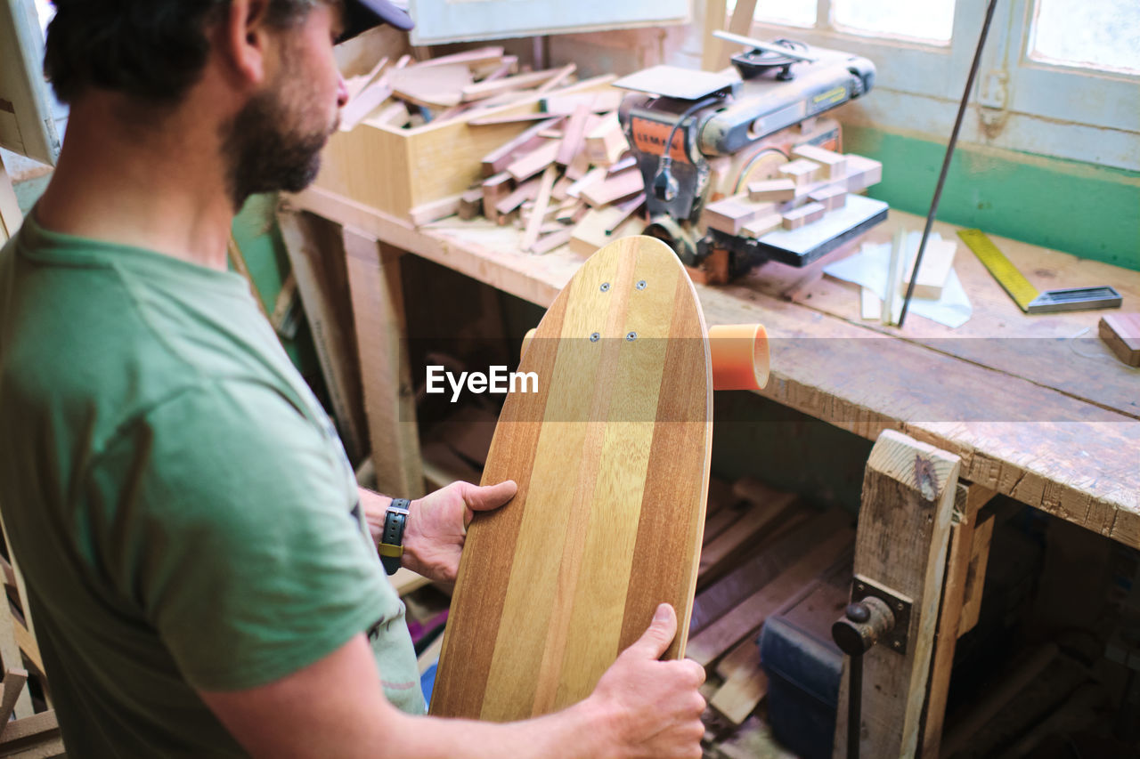 Side view of crop mature bearded male artisan with longboard against professional equipment in workshop