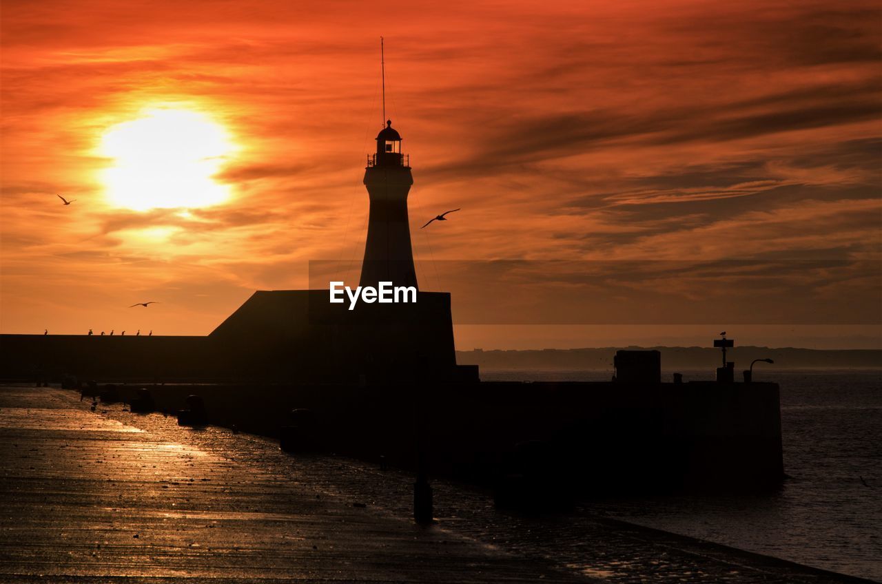 Silhouette lighthouse by sea against orange sky