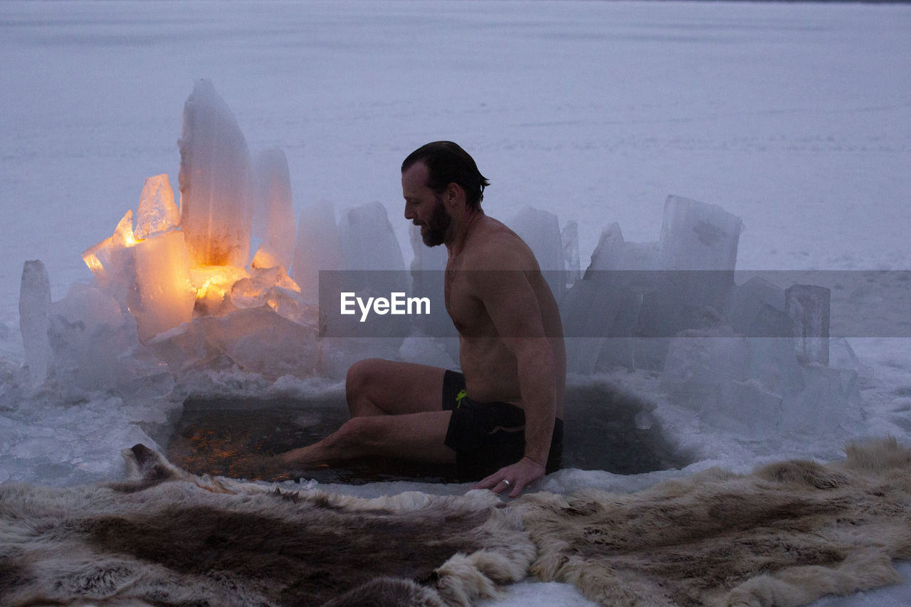 Side view of mature man taking ice bath at frozen lake during dusk