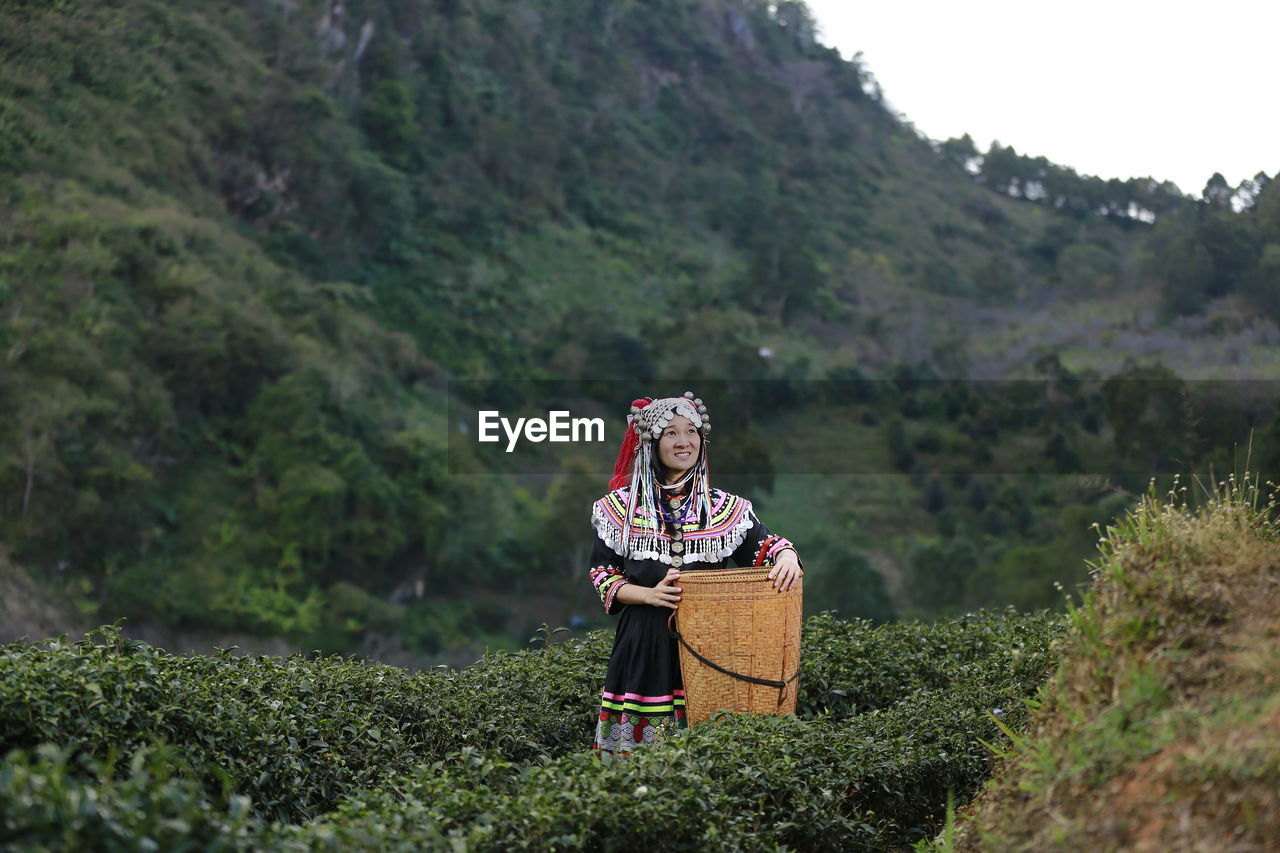 Smiling woman with wicker basket standing amidst tea crops on farm