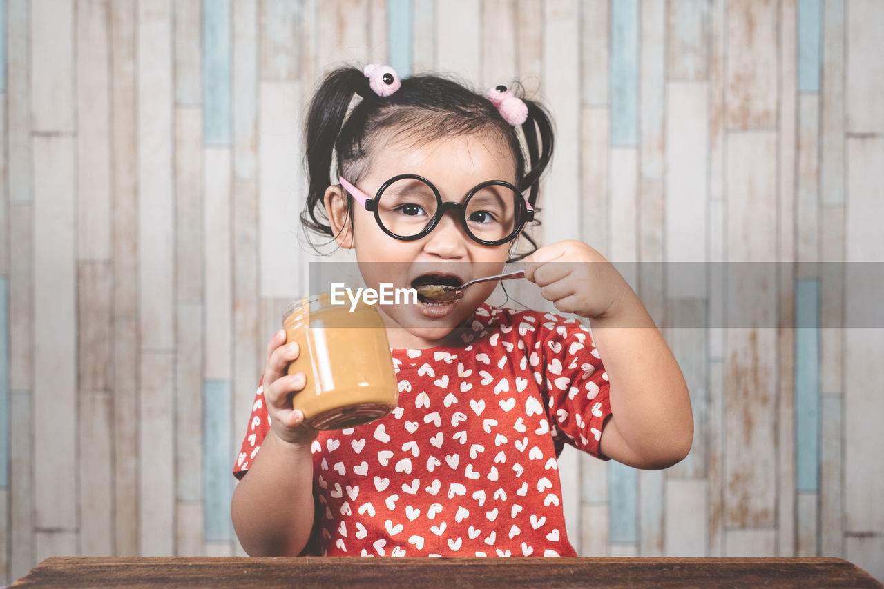 Portrait of girl eating peanut butter at table against wall