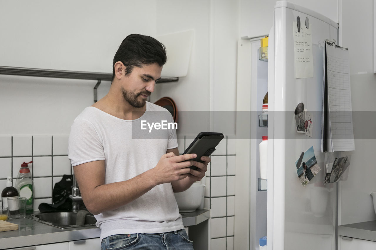 Young man using digital tablet while standing by open refrigerator at kitchen