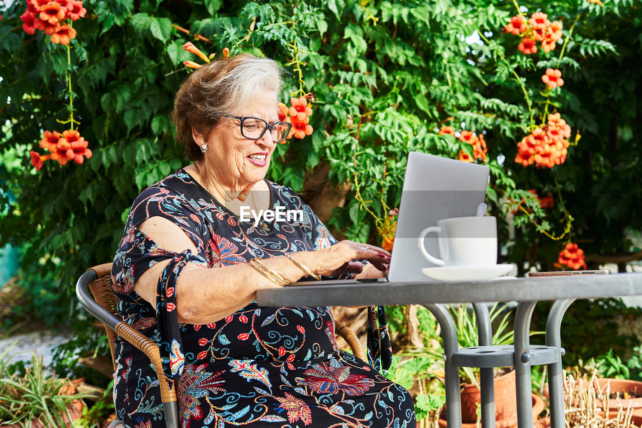 Positive elderly lady in casual outfit and eyeglasses sitting in chair at table while surfing on netbook near cup on saucer and green plants with flowers in yard in daylight