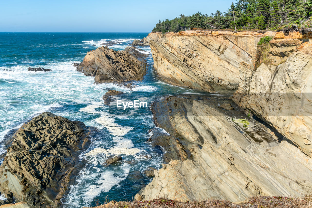 sea, water, rock, land, cliff, beach, coast, shore, beauty in nature, scenics - nature, nature, terrain, sky, body of water, ocean, day, rock formation, no people, horizon over water, wave, geology, horizon, tranquility, tranquil scene, outdoors, motion, coastline, cove, blue, travel destinations, bay, non-urban scene, vacation, sports, idyllic, clear sky, travel, sunlight, water sports, rocky coastline, environment, tree