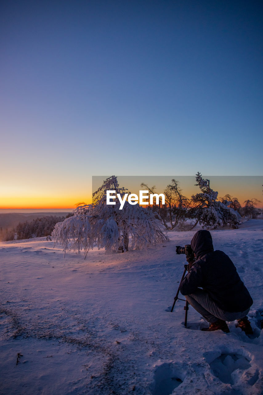 Man taking a photo on snow covered landscape against clear sky during sunset